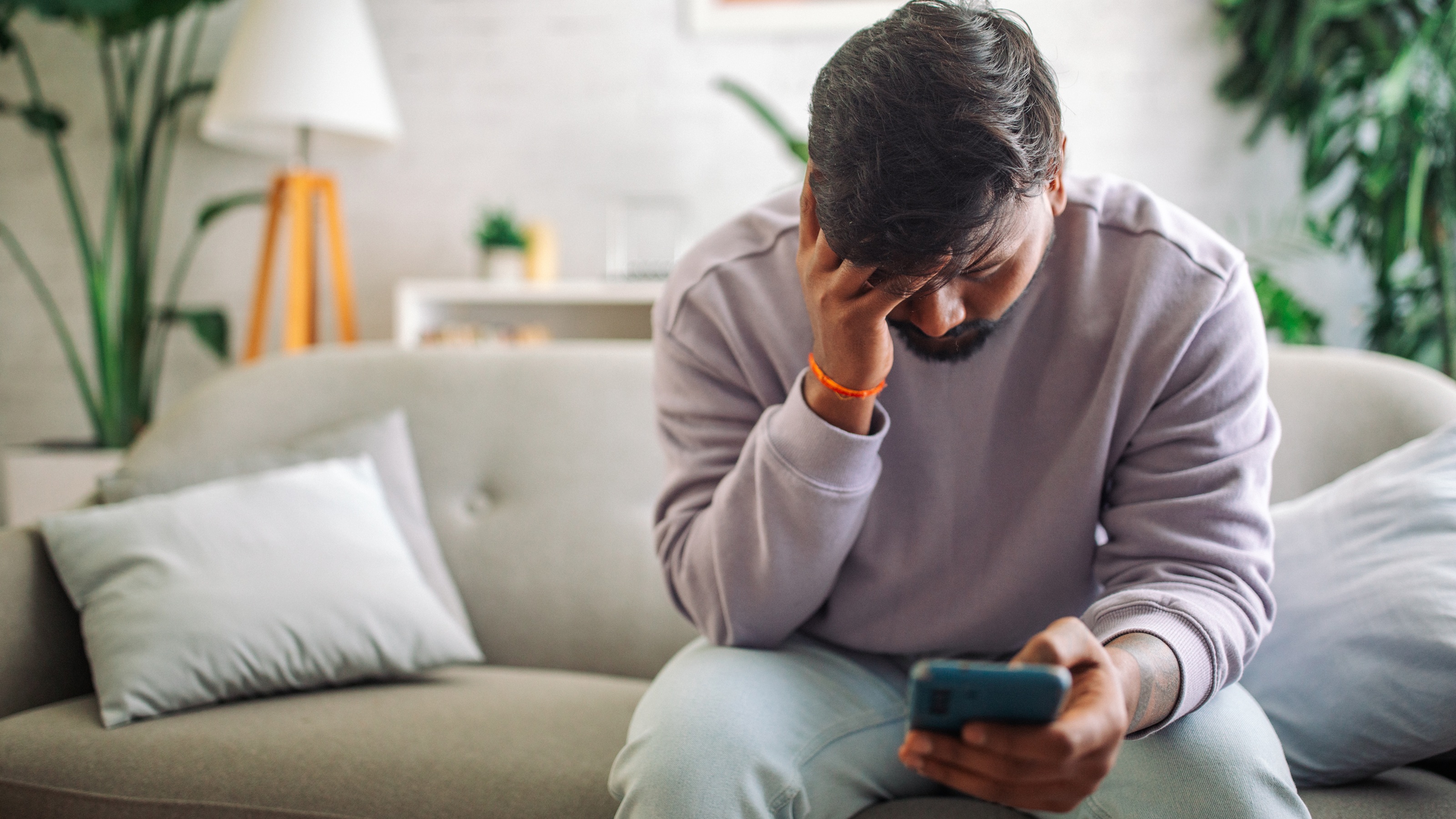 A man appears dejected as he looks at his smartphone while sitting on his sofa.