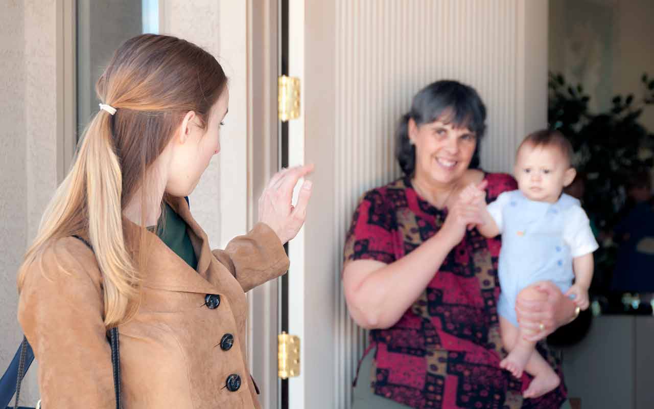 A mother leaving her house and saying goodbye to a nanny who is holding a young child.
