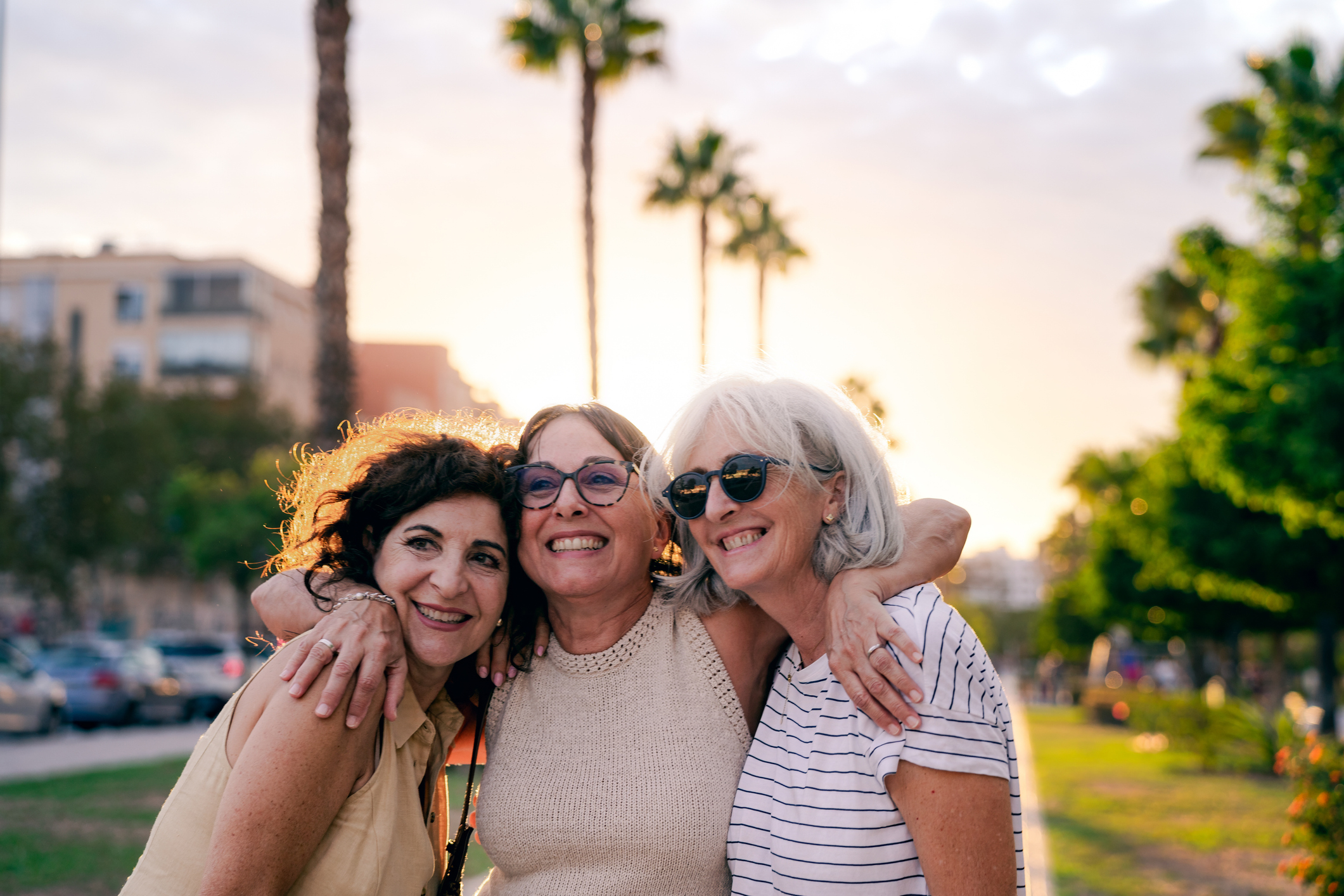 Three elderly women smiling and embracing in the sunset light on a city street.