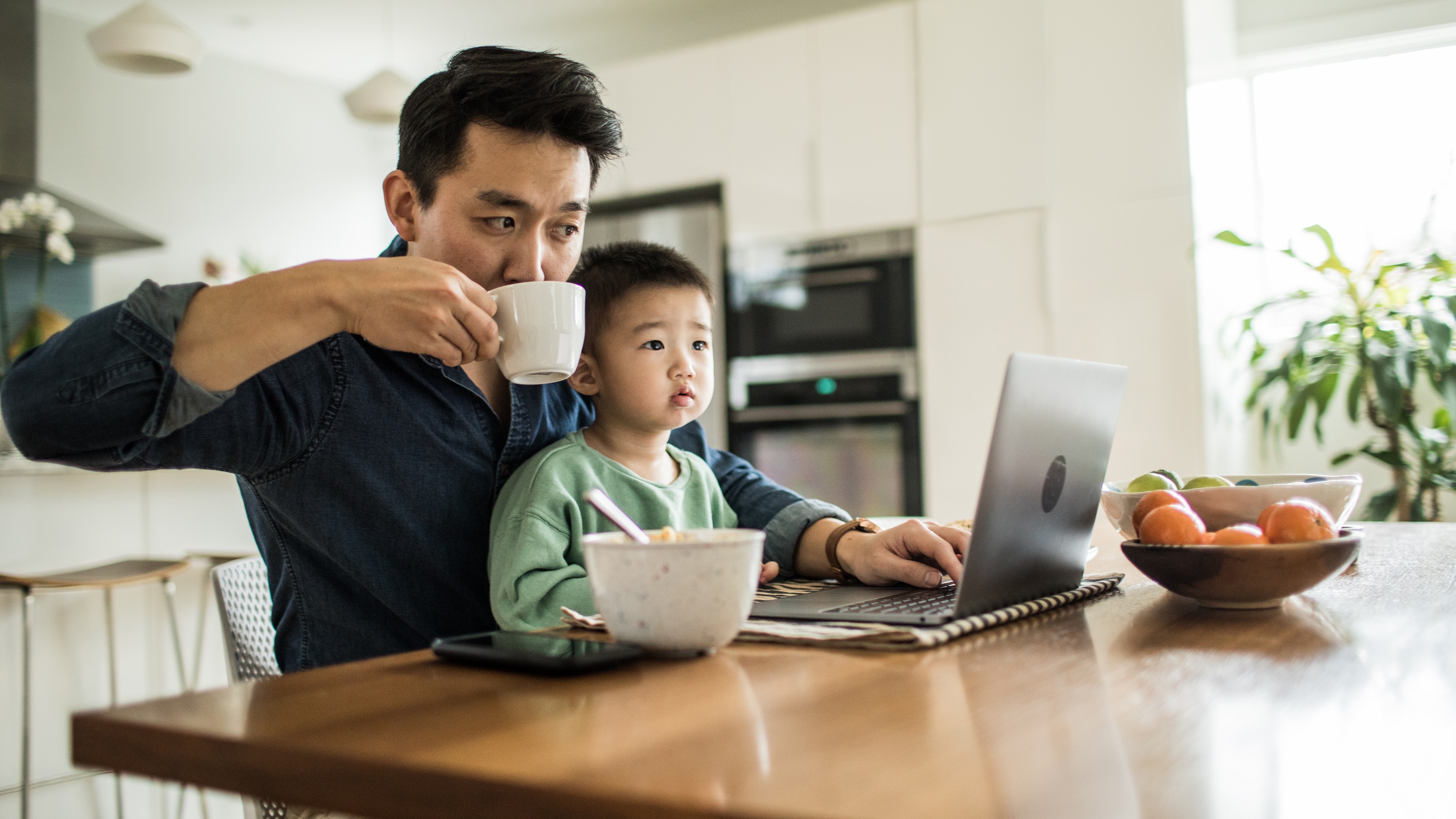 A father works on his laptop at the kitchen table while his young son sits on his lap.