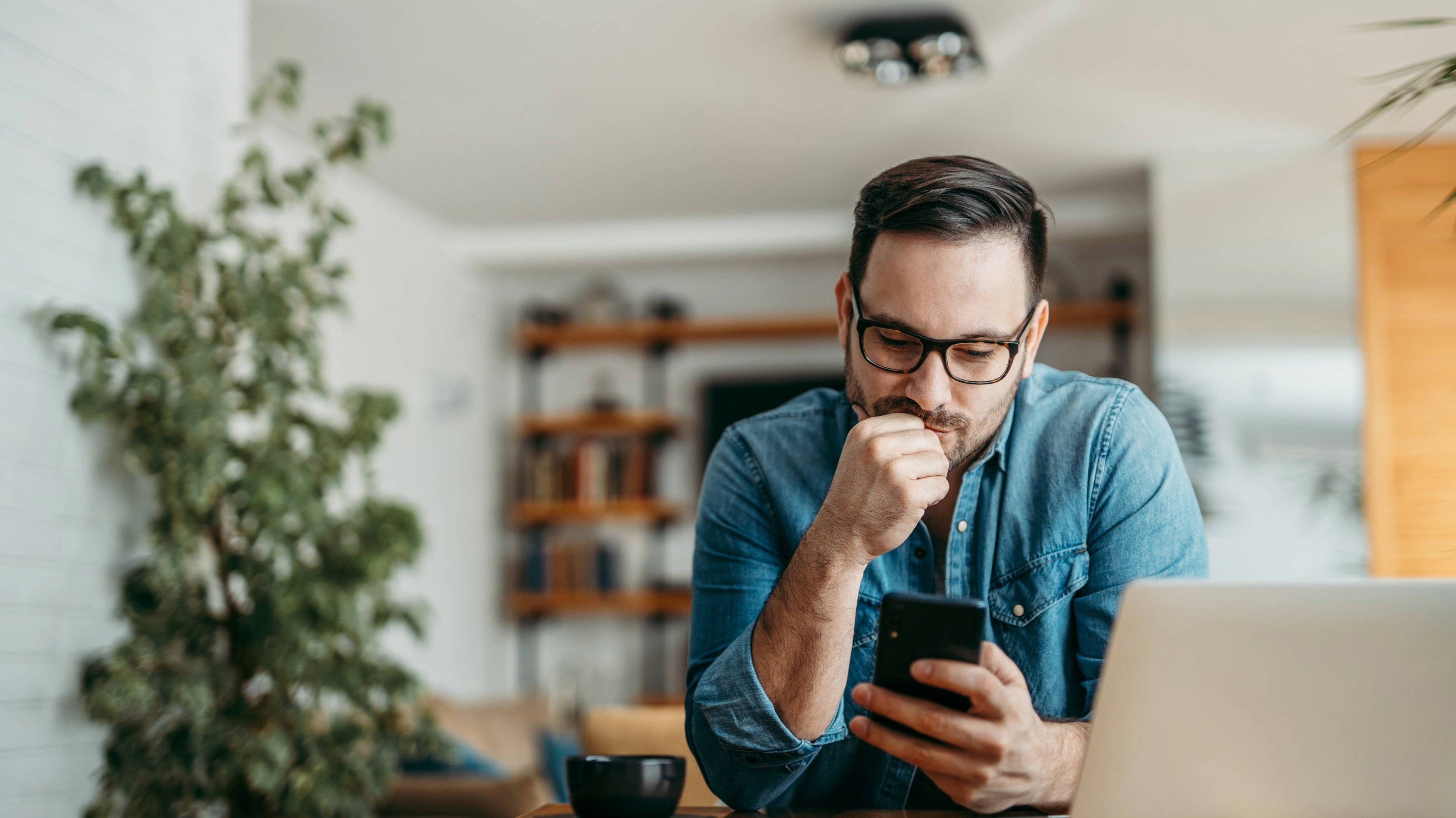 A pensive man sitting at home looking at his smartphone, reflecting the concern over market fluctuations.